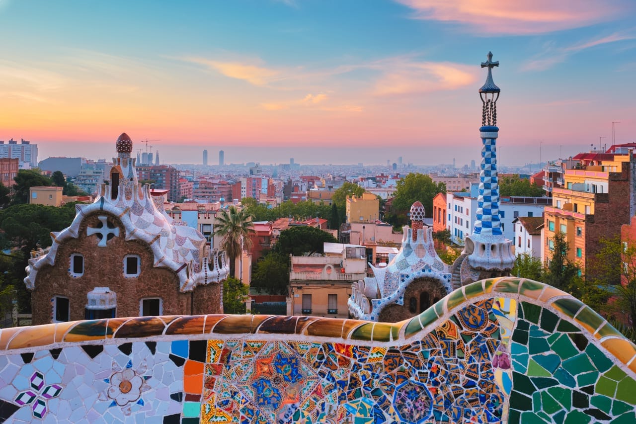 Blick über die Mosaikbank und die Gaudí-Pavillons des Park Güell auf Barcelona im Sonnenaufgang