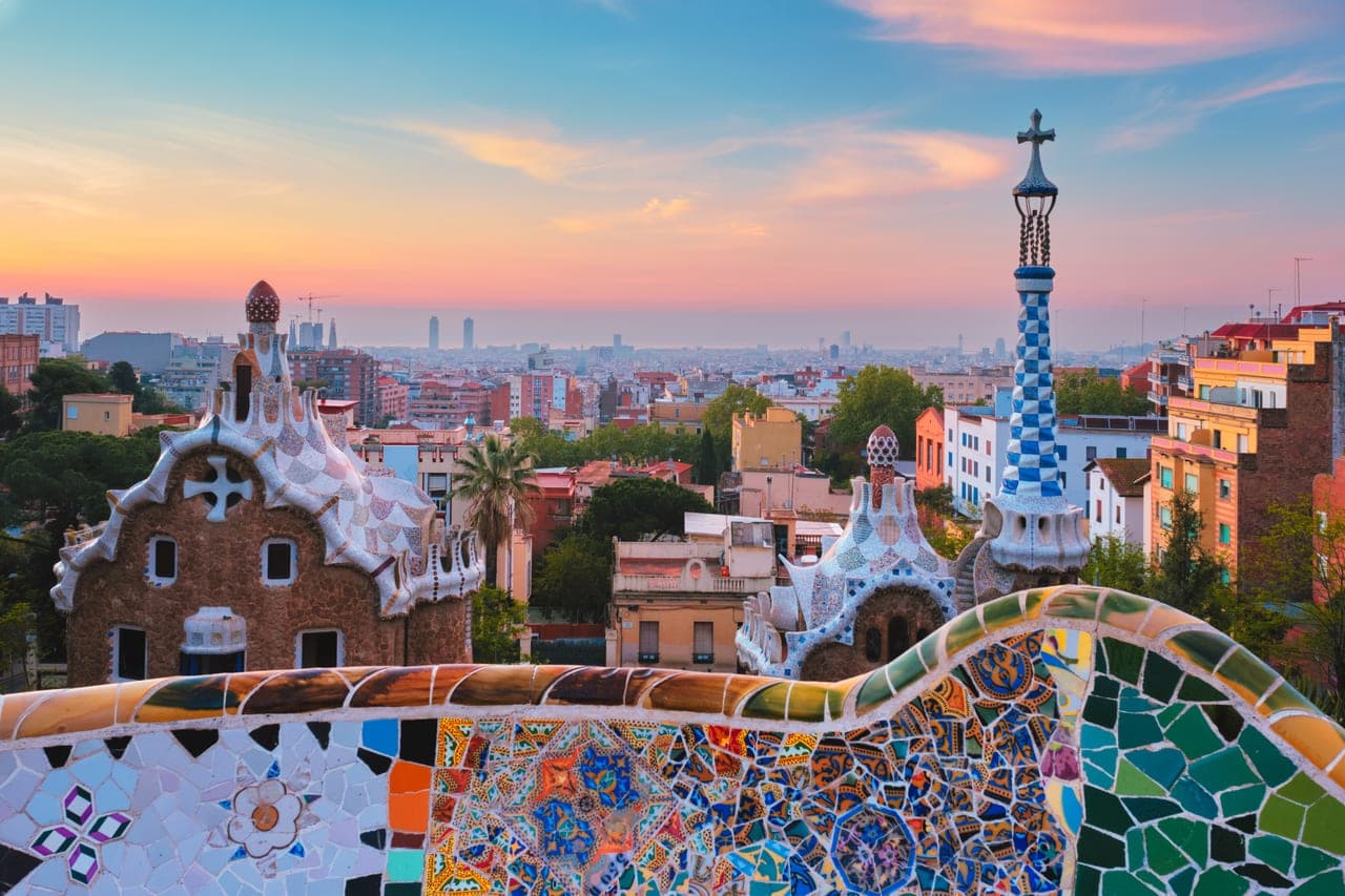 Blick über die Mosaikbank und die Gaudí-Pavillons des Park Güell auf Barcelona im Sonnenaufgang