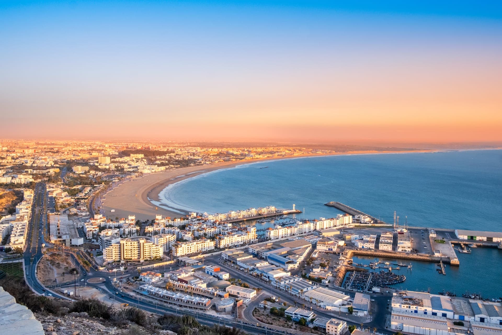 Blick von oben über die geschwungene Bucht von Agadir mit langem Sandstrand, Hafen und der weißen Stadt im Abendlicht