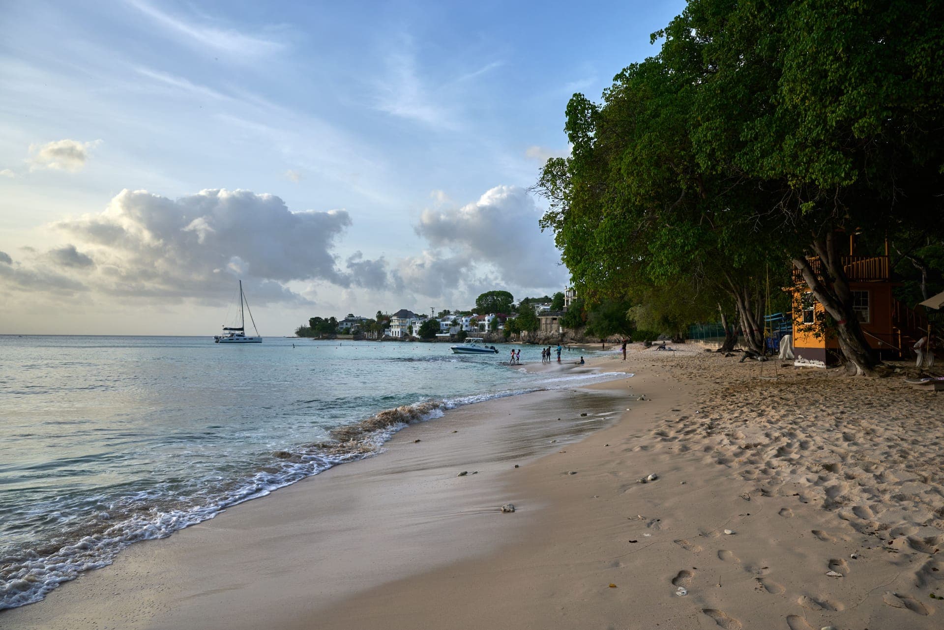 Sandstrand an der Westküste von Barbados im Abendlicht, mit einer auslaufenden Welle, einer ankernden Segelyacht und Bäumen, die bis an den Sand reichen