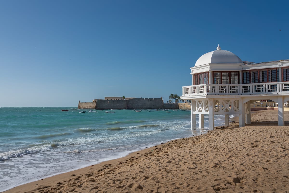 Die Playa La Caleta in Cádiz mit dem weißen Balneario-Pavillon auf Stelzen rechts und dem Castillo de Santa Catalina auf der gegenüberliegenden Landzunge