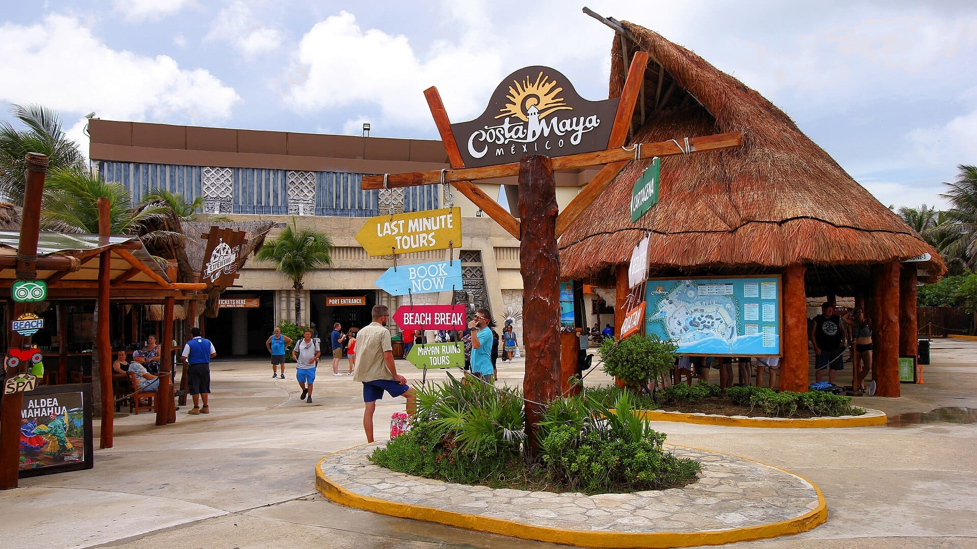 Der Vorplatz am Ausgang des Hafendorfs von Costa Maya mit großem Schriftzug „Costa Maya México", einem palmwedelgedeckten Rundpavillon, Wegweisern zu Ausflügen und Kreuzfahrtgästen