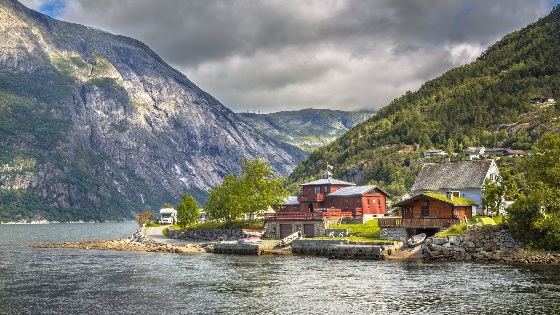 Rote und weiße Holzhäuser auf einer Landzunge am inneren Hardangerfjord, dahinter steil aufragende Bergflanken unter Wolken