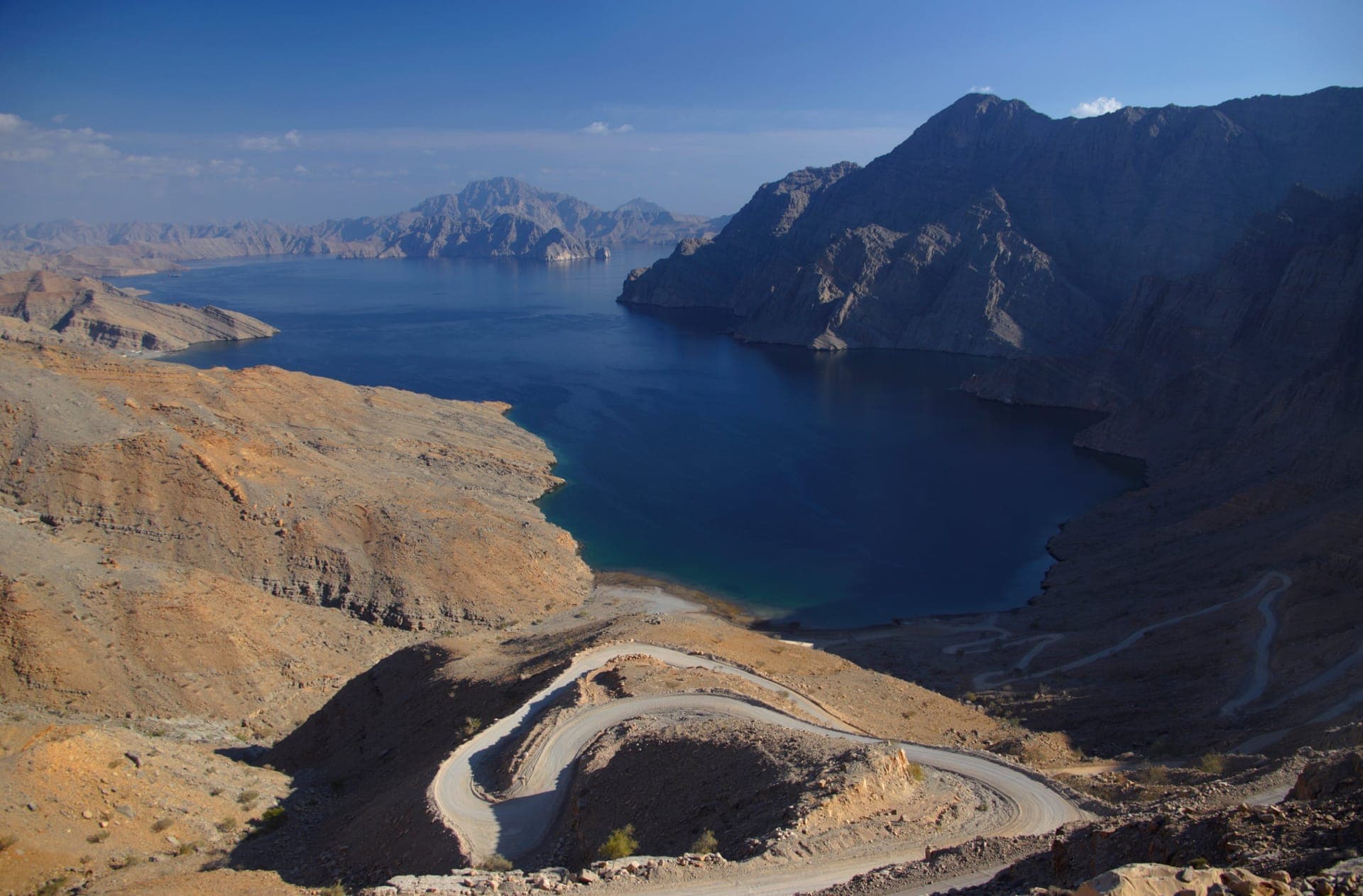Blick über einen der Musandam-Fjorde bei Khasab mit tiefblauem Wasser zwischen kargen Felswänden, im Vordergrund eine sich windende Schotterpiste