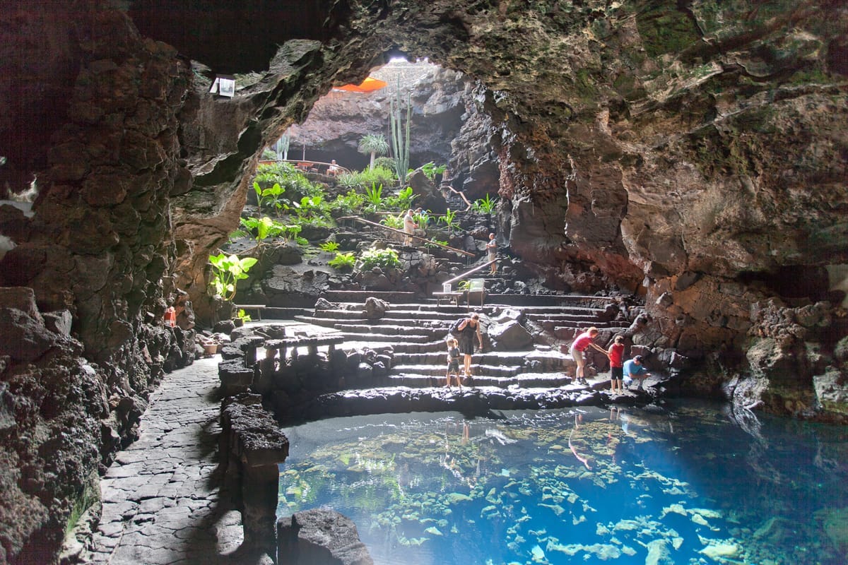 Blick aus der Lavahöhle Jameos del Agua auf den türkis leuchtenden unterirdischen See, dahinter eine Treppe hinauf zur offenen, bepflanzten Höhlendecke