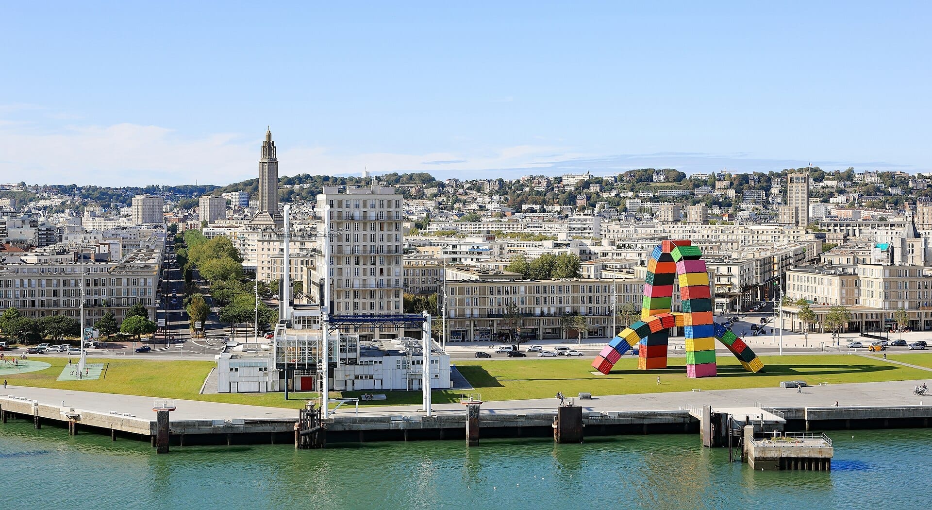 Blick vom Wasser auf Le Havre mit dem Betonturm der Kirche St-Joseph links, der hellen Perret-Nachkriegsbebauung und der bunten Container-Skulptur Catène de Containers am Ufer