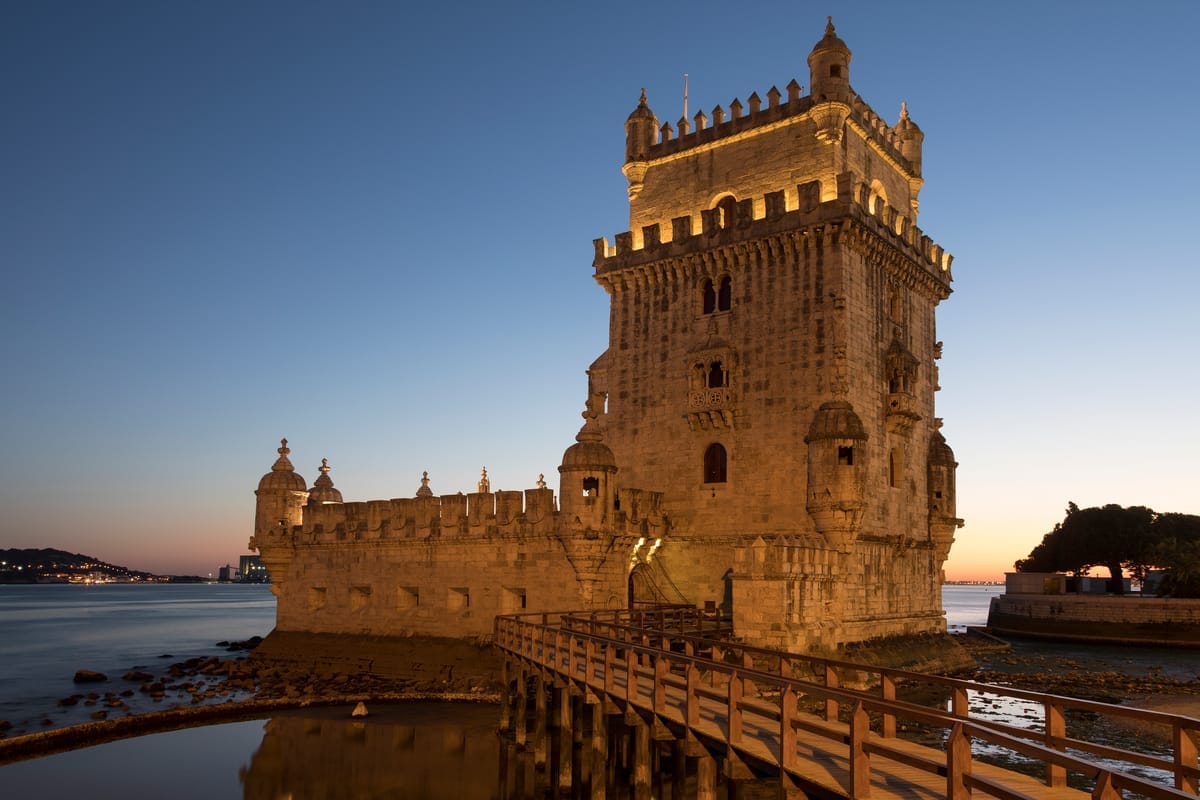 Der Torre de Belém am Ufer des Tejo in Lissabon, warm angestrahlt vor blauem Abendhimmel, davor der hölzerne Zugangssteg