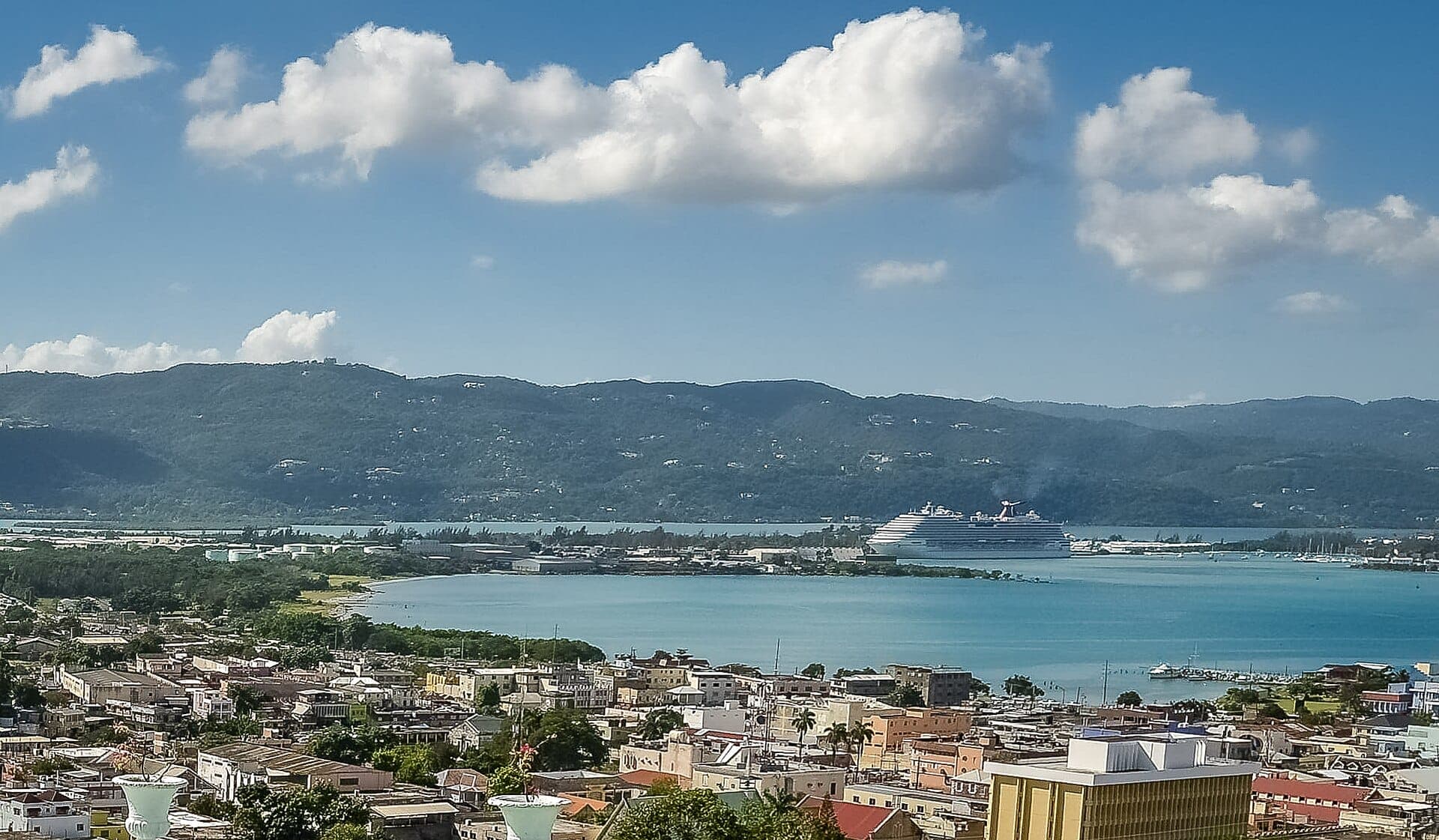 Blick von einem Hügel über die Dächer von Montego Bay auf die türkisfarbene Bucht, ein Kreuzfahrtschiff am Terminal und die bewaldeten Berge dahinter