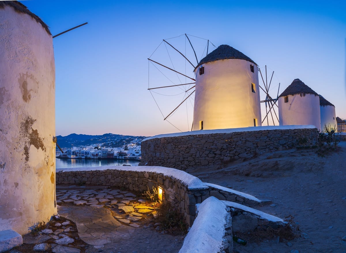 Die Windmühlen von Kato Mili in der Abenddämmerung, im Hintergrund die beleuchteten Häuser von Klein-Venedig am Wasser
