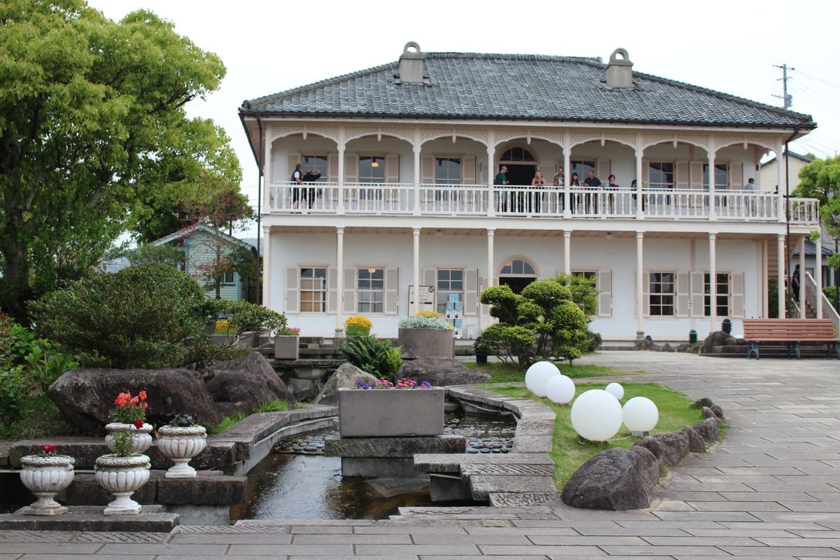 Weiße Kolonialvilla mit umlaufender Veranda im Glover Garden in Nagasaki, davor ein kleiner Wasserlauf und Blumenkübel