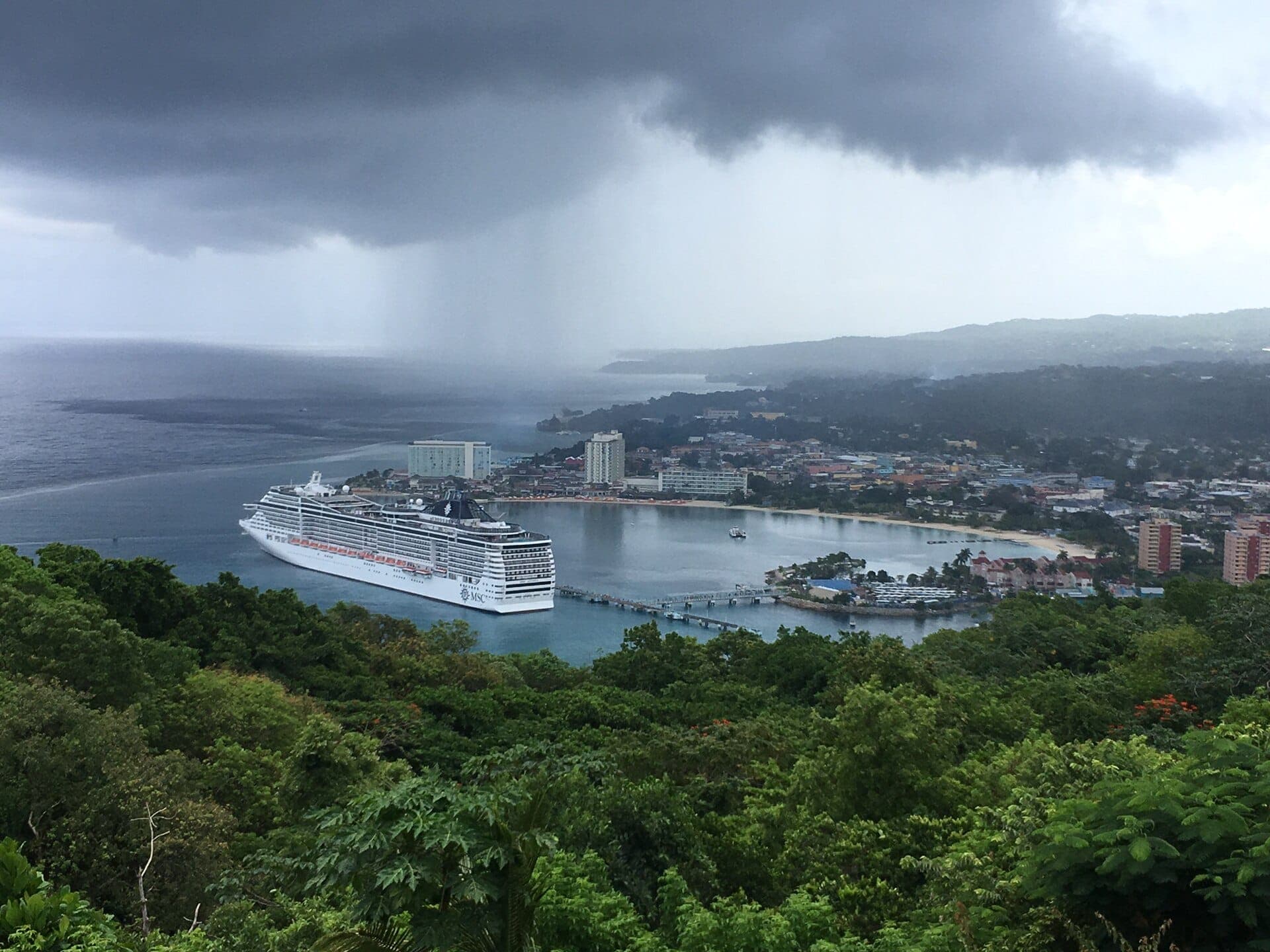 Blick von einem bewaldeten Hügel auf die Bucht von Ocho Rios mit einem Kreuzfahrtschiff am Pier, dem Stadtstrand, den Hotels der Stadt und einem Regenschauer über dem Meer