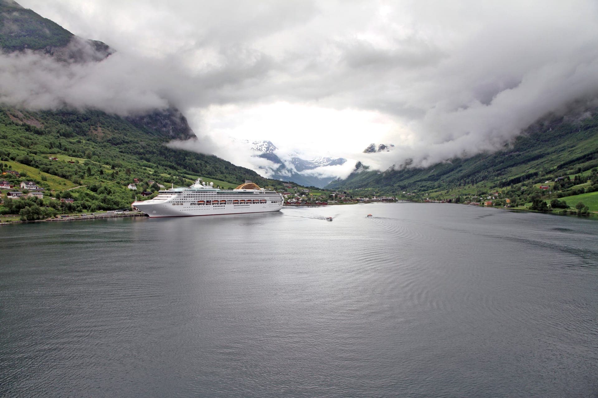 Kreuzfahrtschiff im Nordfjord vor dem Ort Olden, die grünen Berghänge in tief hängenden Wolken, dahinter schneebedeckte Gipfel