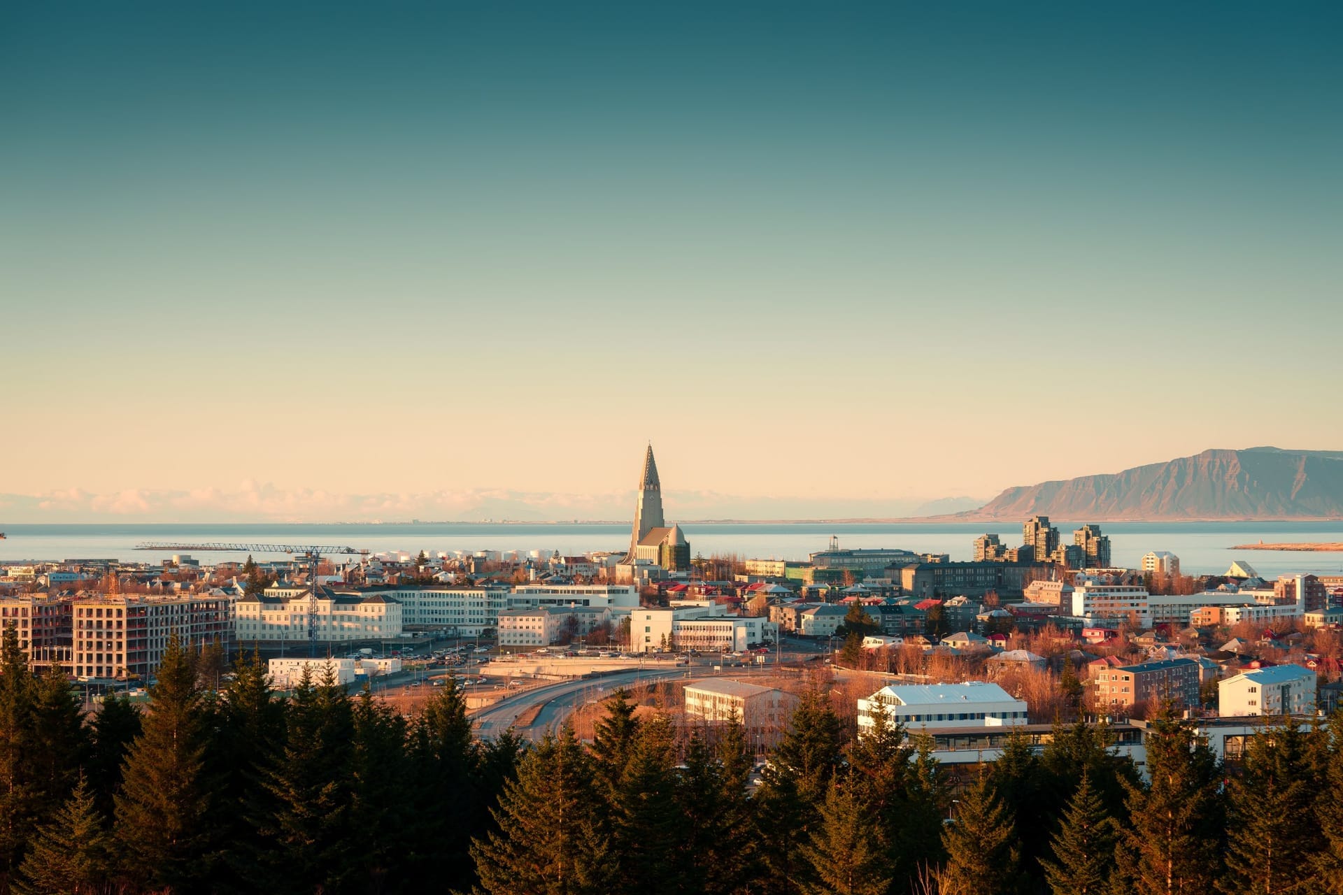 Blick über Reykjavik im Abendlicht mit dem Turm der Hallgrímskirkja über den Dächern, dahinter die Bucht und die Berge
