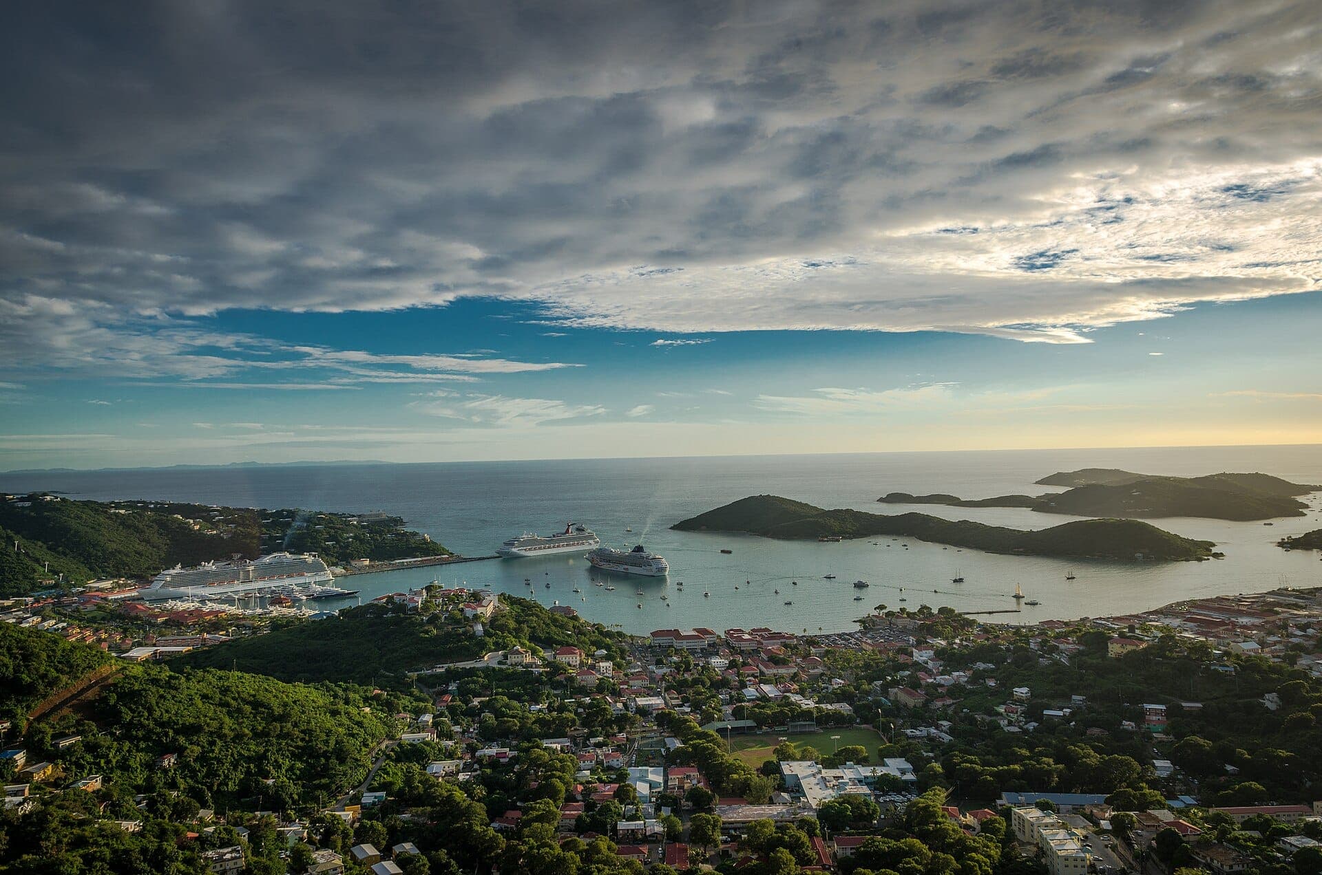 Blick von den Hügeln über die Dächer von Charlotte Amalie auf die weit geschwungene Hafenbucht mit mehreren Kreuzfahrtschiffen, ankernden Yachten und vorgelagerten grünen Inseln im Abendlicht