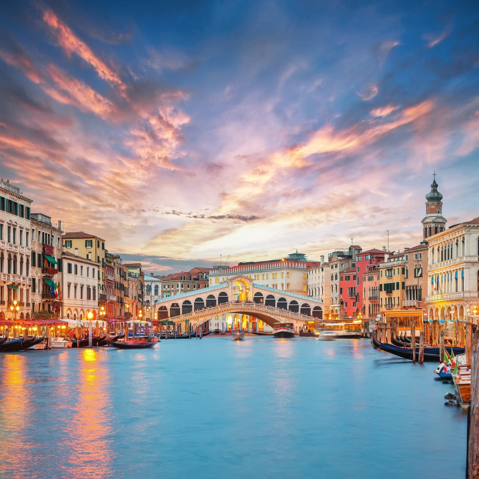 Die Rialtobrücke über dem Canal Grande im Abendlicht, davor Gondeln und beleuchtete Palazzi