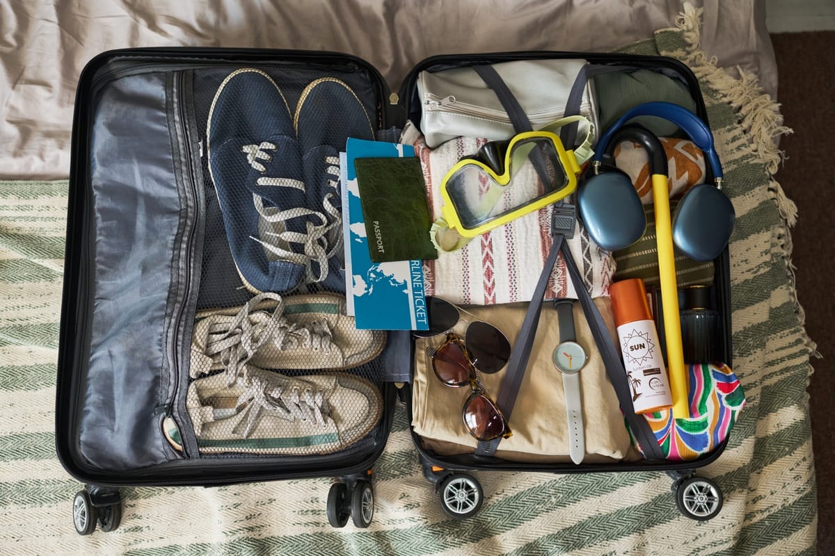 Geöffneter, ordentlich gepackter Koffer auf einem Bett, darin Schuhe, Reisepass, Ticket, Sonnenbrille, Sonnencreme und Kopfhörer