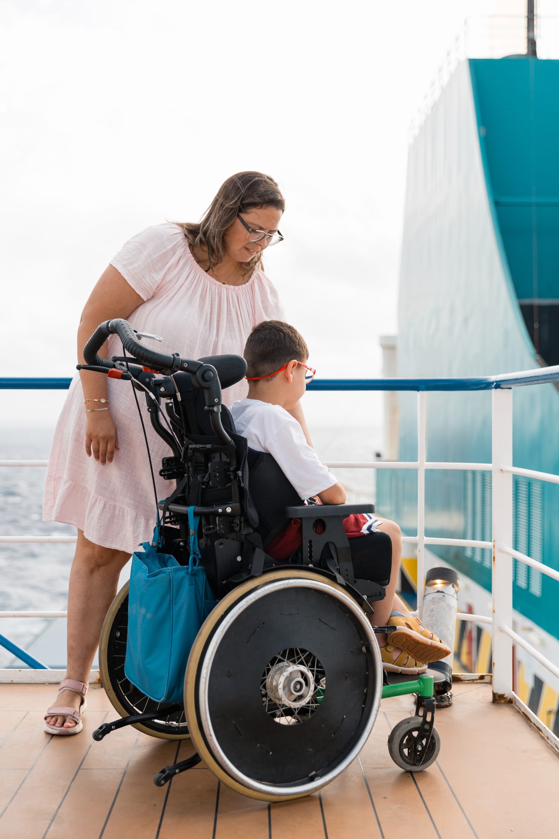 Eine Frau steht auf einem sonnigen Schiffsdeck neben einem Kind im Rollstuhl, dahinter die Reling und das Meer