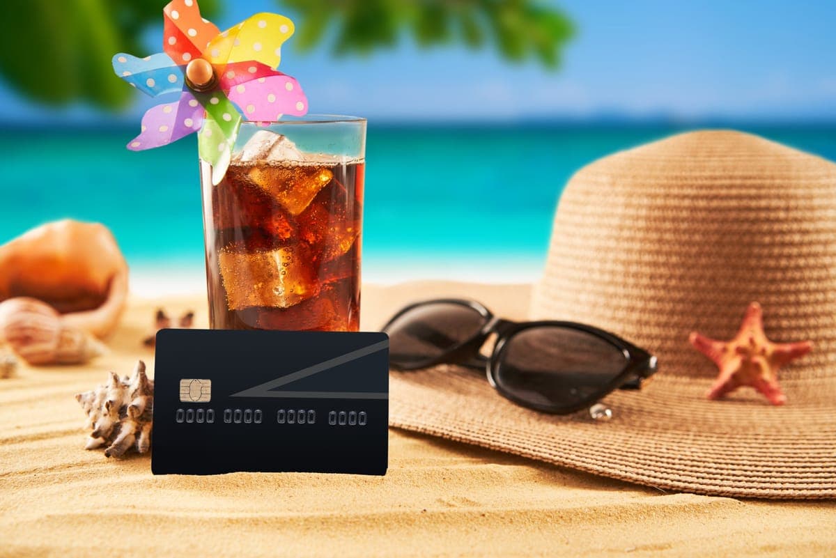 Kreditkarte im Sand zwischen Sonnenhut, Sonnenbrille und einem Cocktail am Strand