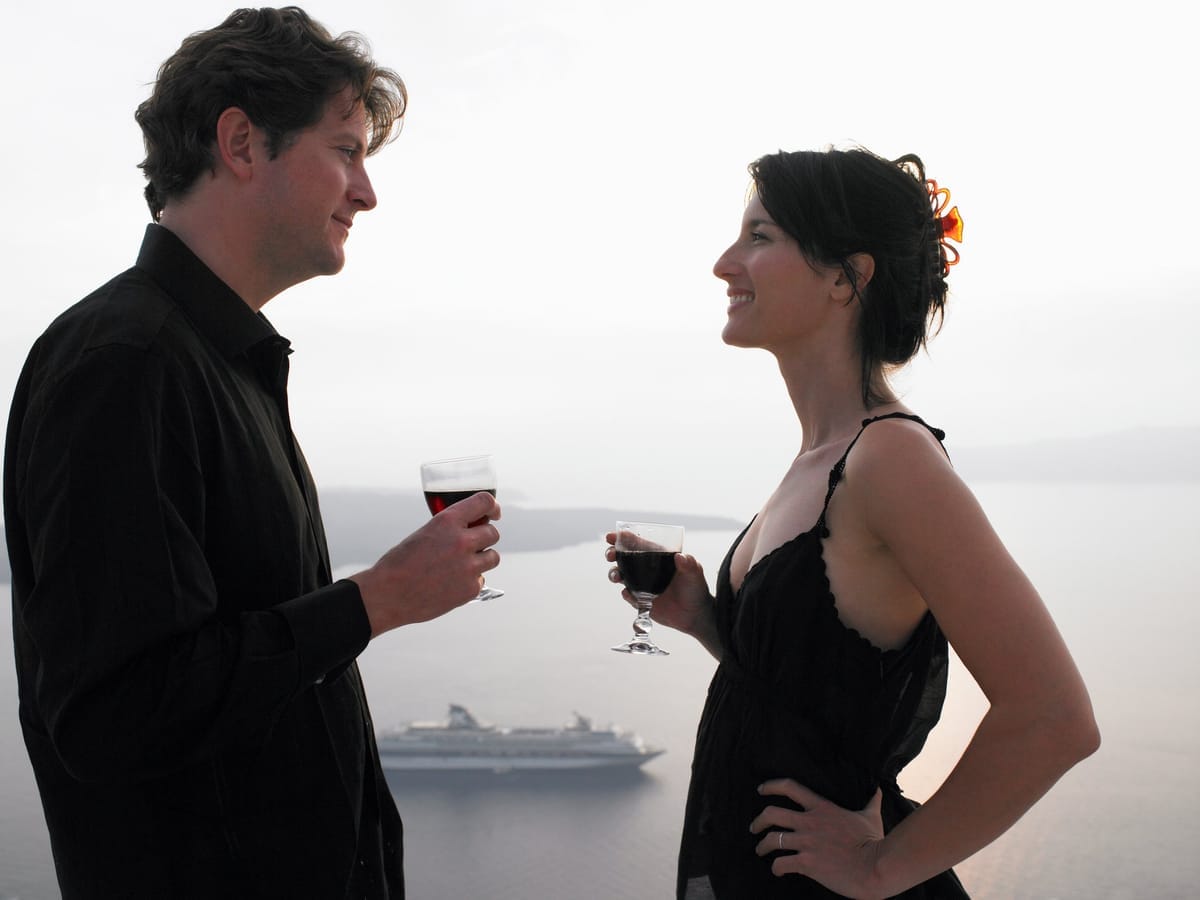 Elegant gekleidetes Paar stößt mit Weingläsern an, im Hintergrund ein Kreuzfahrtschiff bei Sonnenuntergang