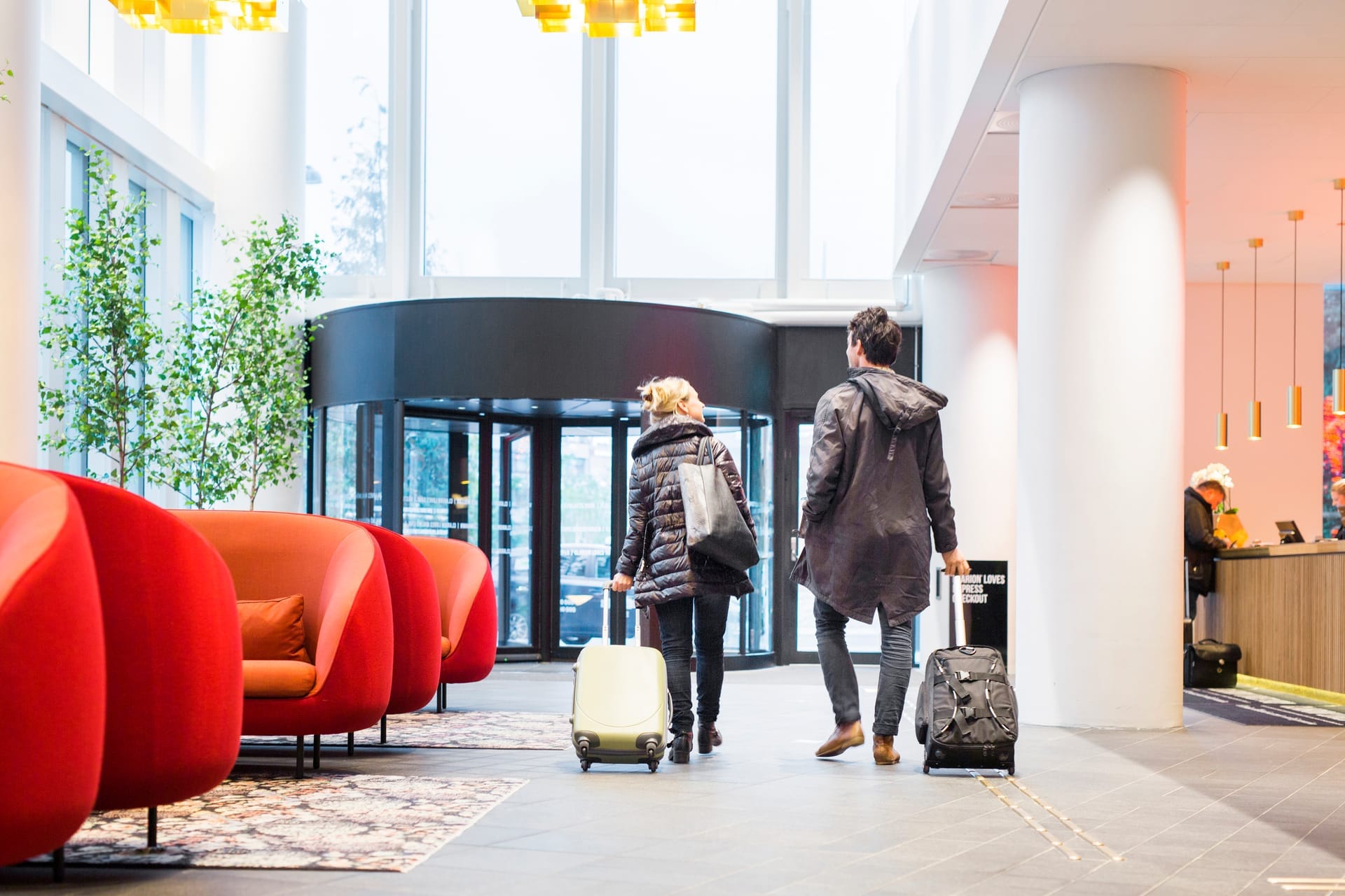 Zwei Reisende ziehen ihre Rollkoffer durch eine helle Hotellobby Richtung Drehtür, rechts die Rezeption