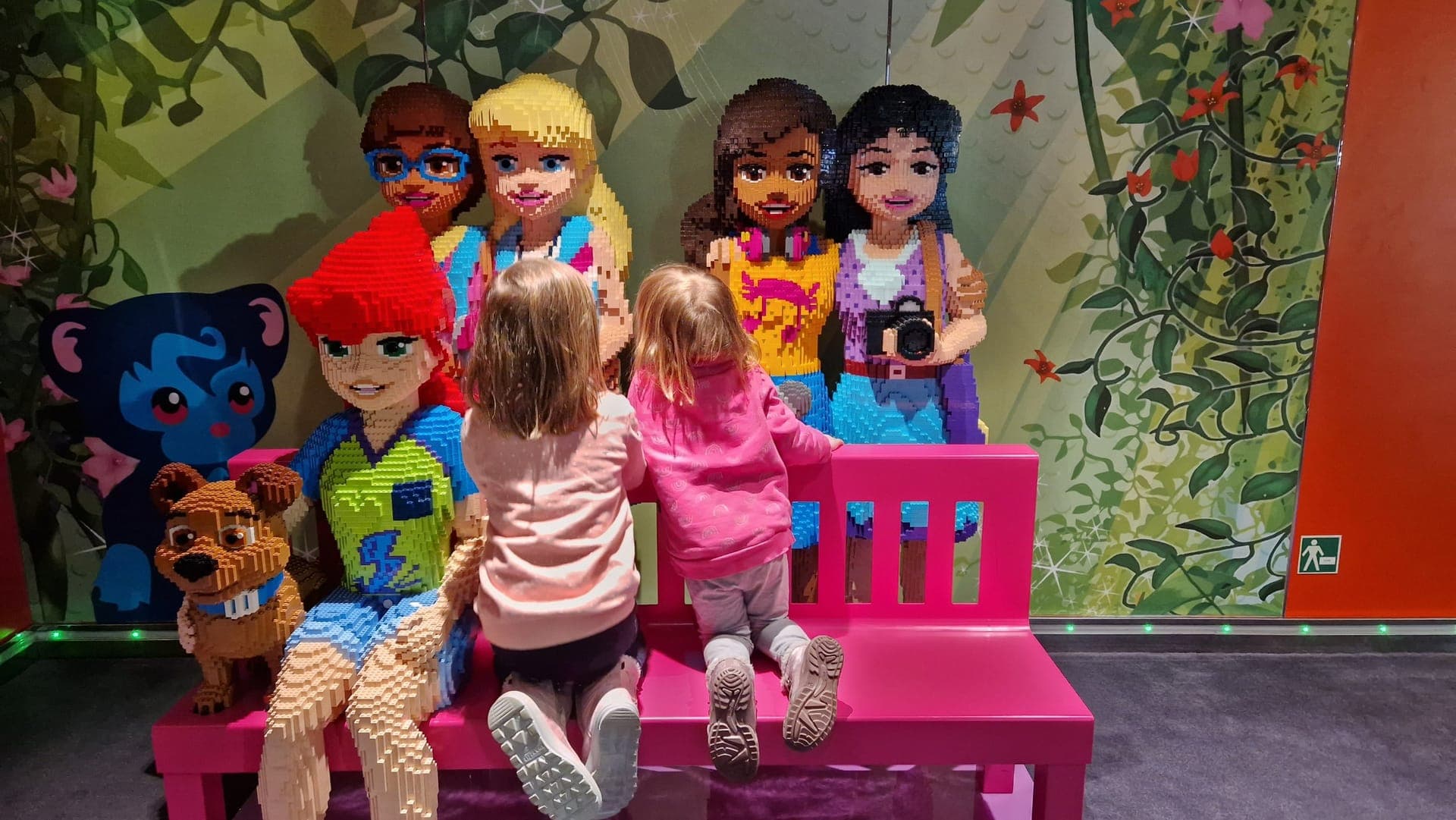Zwei kleine Kinder knien von hinten fotografiert auf einer pinken Bank im Kinderbereich eines Kreuzfahrtschiffs und schauen auf lebensgroße Figuren aus Bausteinen vor einer bunt bemalten Dschungelwand