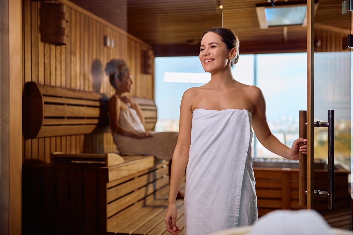 Frau in Handtuch verlässt lächelnd eine Holzsauna, im Hintergrund eine weitere Person auf der Sitzbank