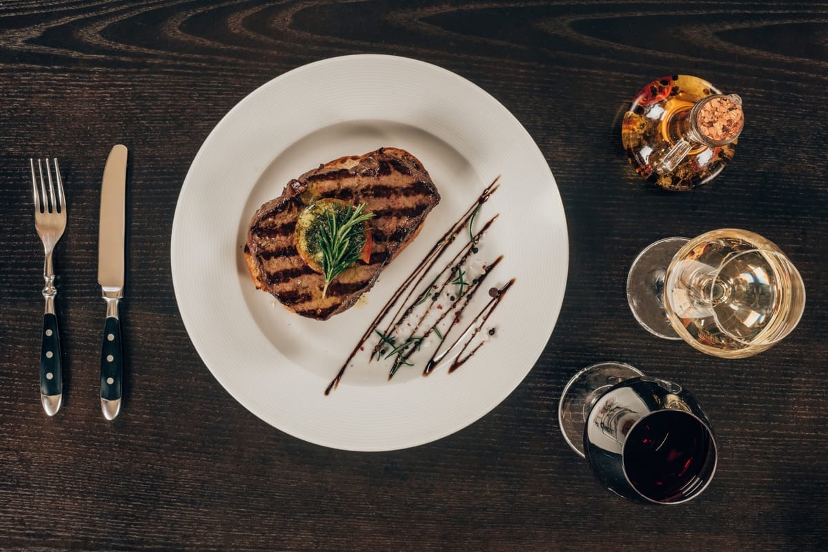 Gegrilltes Steak auf einem Teller, daneben Wein- und Rotweinglas auf dunklem Holztisch