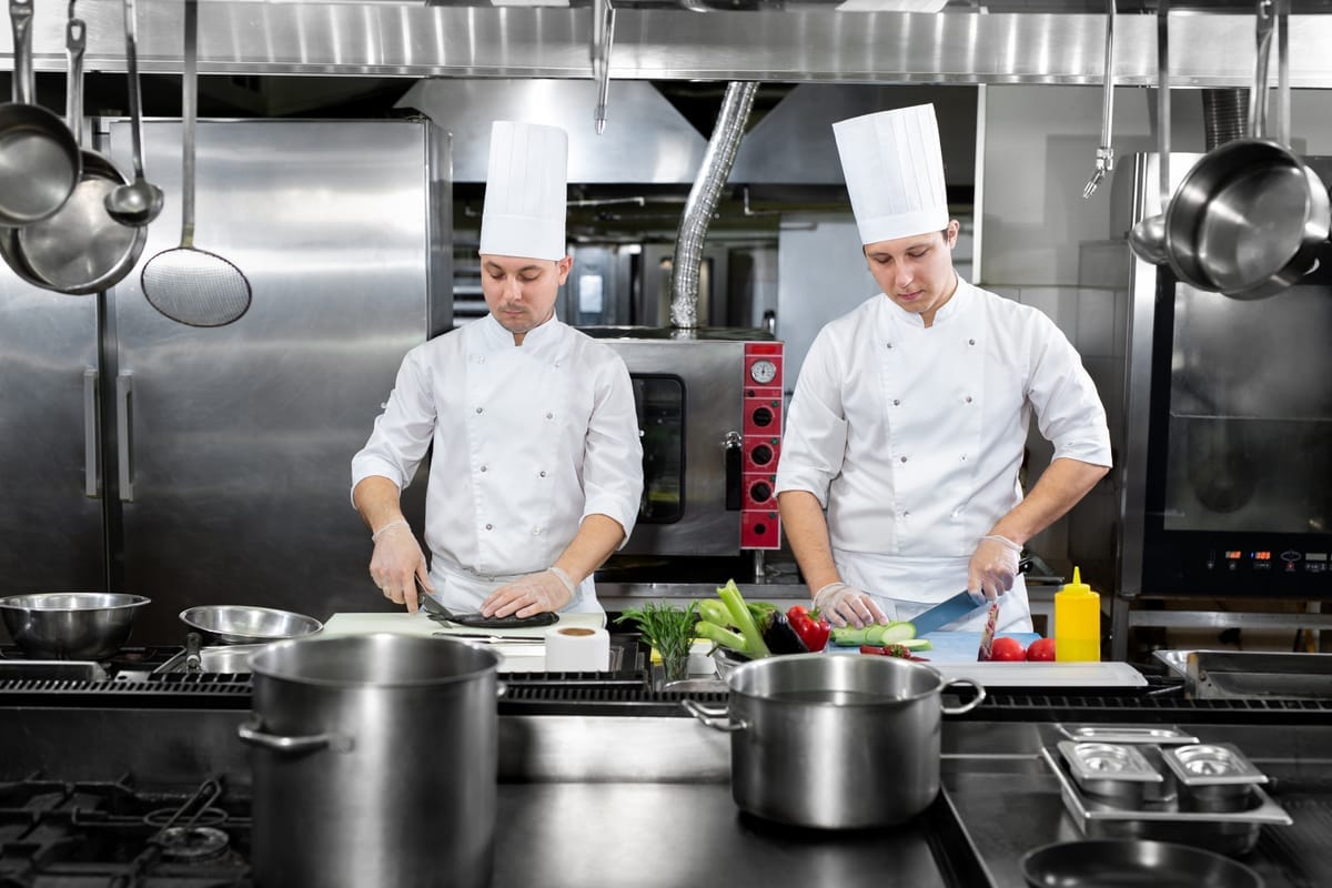 Zwei Köche in weißer Uniform schneiden Gemüse an einer Edelstahl-Arbeitsstation in einer Großküche