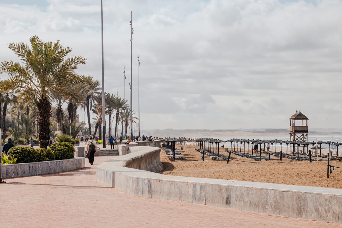Die palmengesäumte Strandpromenade von Agadir, rechts der breite Sandstrand mit zusammengeklappten Sonnenschirmen und ein hölzerner Rettungsschwimmerturm