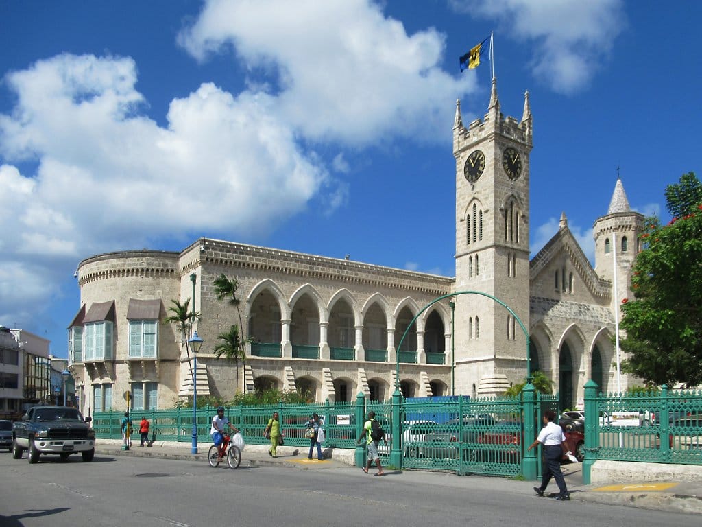 Die neugotischen Parliament Buildings von Bridgetown aus hellem Korallenstein mit Uhrturm und der Flagge von Barbados, davor ein grüner Eisenzaun und Fußgänger