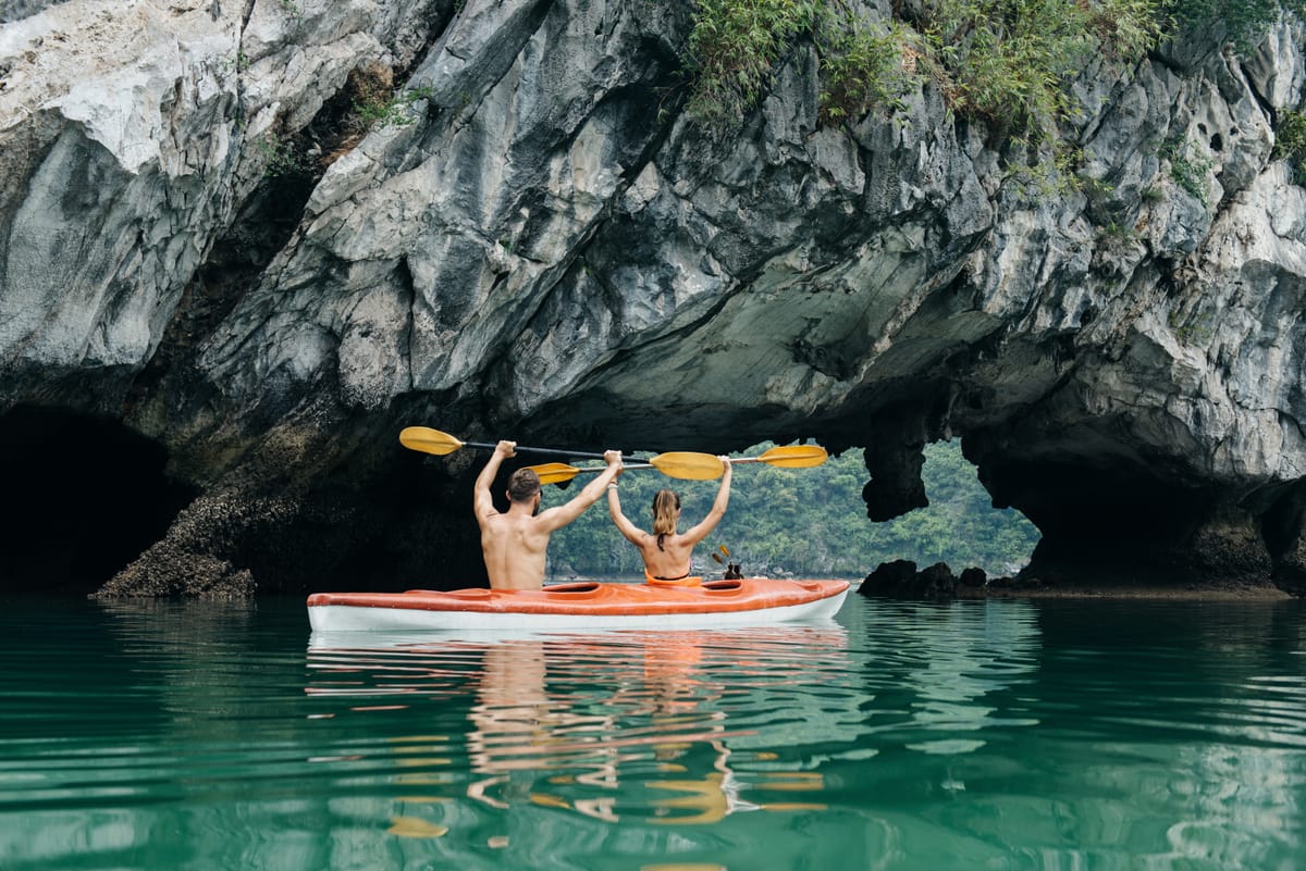 Zwei Personen in einem Kajak vor der Einfahrt einer Felshöhle in der Halong-Bucht, ringsum steile Kalksteinwände