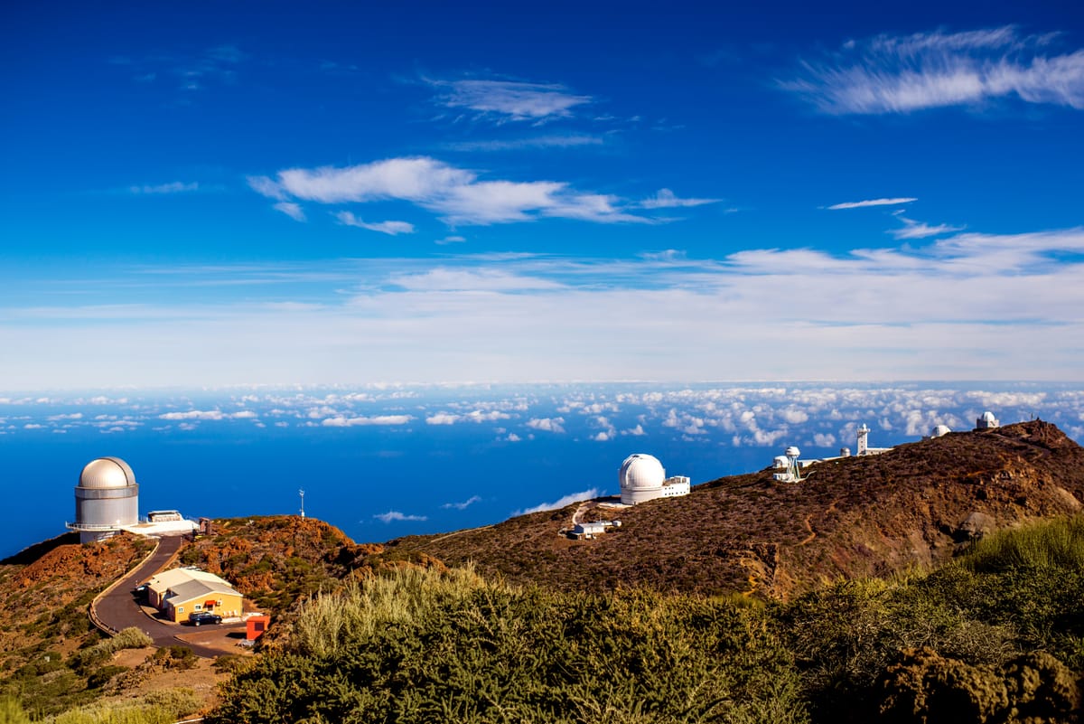 Mehrere weiße Teleskopkuppeln eines Observatoriums auf einem kahlen Bergrücken, darüber ziehende Wolken