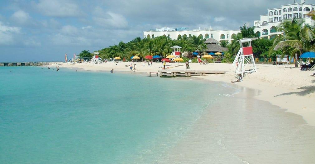 Heller Sandstrand am Doctor's Cave Beach mit flachem, klarem Wasser, bunten Sonnenschirmen, weißem Rettungsschwimmerturm und Hotelgebäuden hinter Palmen