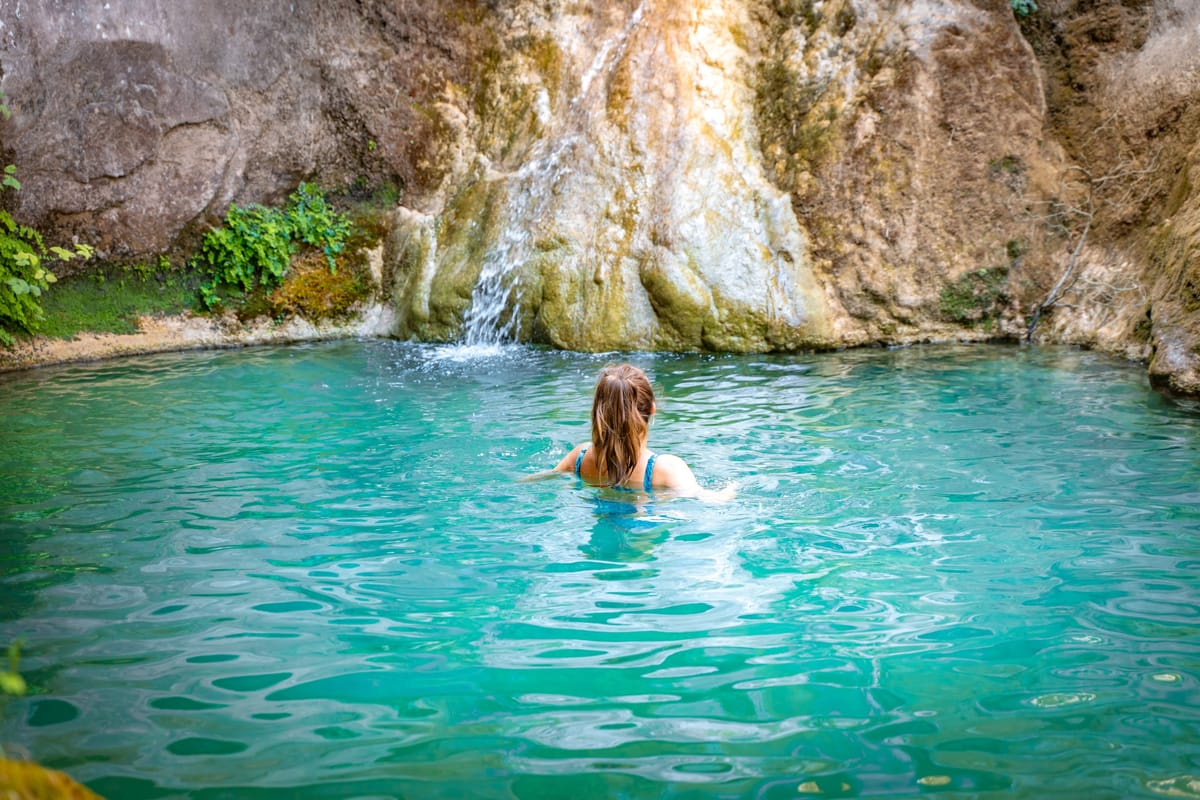 Frau schwimmt in einem türkisblauen Naturpool unter einem kleinen Wasserfall im Dschungel bei Ocho Rios