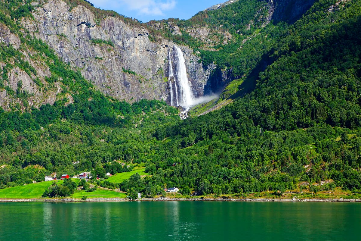 Ein hoher Wasserfall stürzt über eine Felswand in einen grünen Fjordarm bei Skjolden, am Fuß liegt ein kleiner Hof