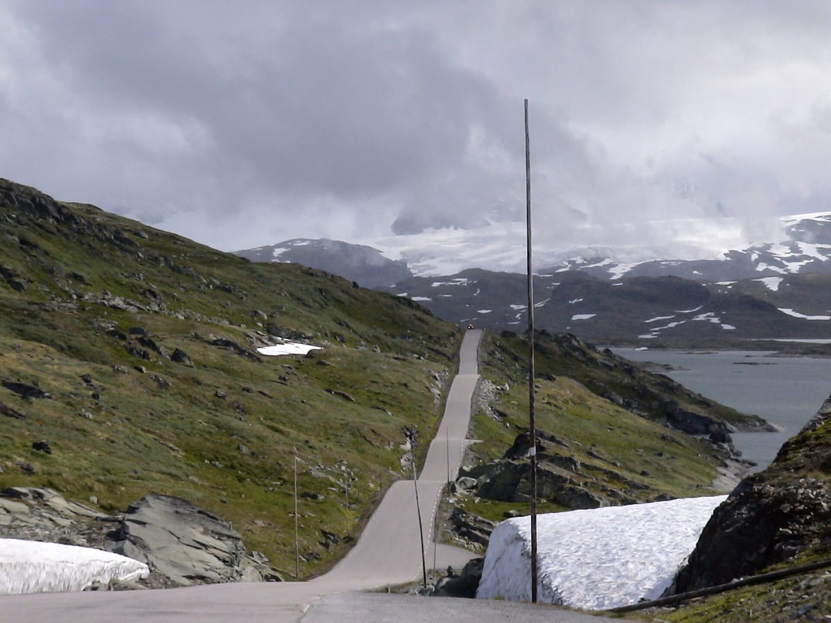 Eine schmale Passstraße läuft über kahles Hochgebirgsplateau auf eine wolkenverhangene Gletscherkette zu, links und rechts der Fahrbahn liegen Schneefelder und ein grau-grüner Gebirgssee