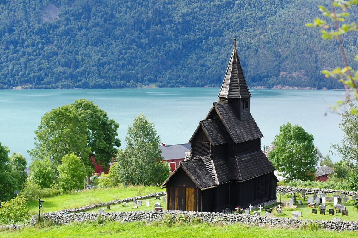 Die dunkel geteerte Stabkirche Urnes mit gestaffelten Schindeldächern und schlankem Dachreiter, umgeben von einem Friedhof mit Trockenmauer, dahinter der Fjord und ein bewaldeter Steilhang