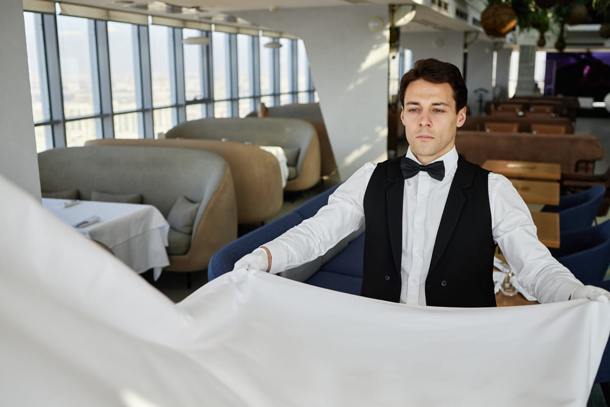 Kellner in Weste und Fliege deckt in einem eleganten Restaurant den Tisch mit einer weißen Tischdecke ein