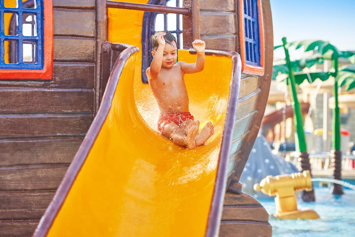 Ein kleiner Junge rutscht mit erhobenen Armen eine gelbe Wasserrutsche hinunter, dahinter der Wasserspielplatz mit Piratenschiff-Aufbauten
