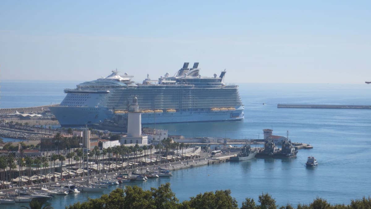Blick von einem Hügel auf einen Mittelmeerhafen: Ein Kreuzfahrtschiff der Oasis-Klasse liegt an der Mole und überragt Leuchtturm, Promenade, Segelboote und Schlepper um ein Vielfaches