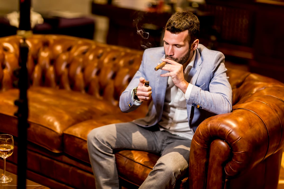 Ein Mann sitzt auf einem Sofa und zündet sich eine Zigarre an, vor ihm steht ein Glas Weißwein